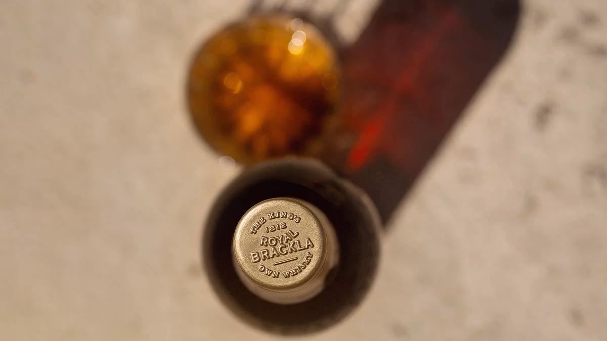 Close-up of Royal Brackla whisky bottle cap with embossed gold lettering and blurred glass of amber liquid.