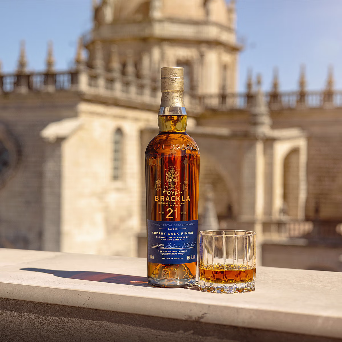 Bottle of Royal Brackla 21 Year Highland Single Malt Scotch Whisky with blue label and gold cap beside cut-glass tumbler.